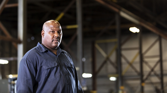 A mature African-American man with a large human build standing in a dimly lit manufacturing warehouse. He is looking away with a serious expression on his face.